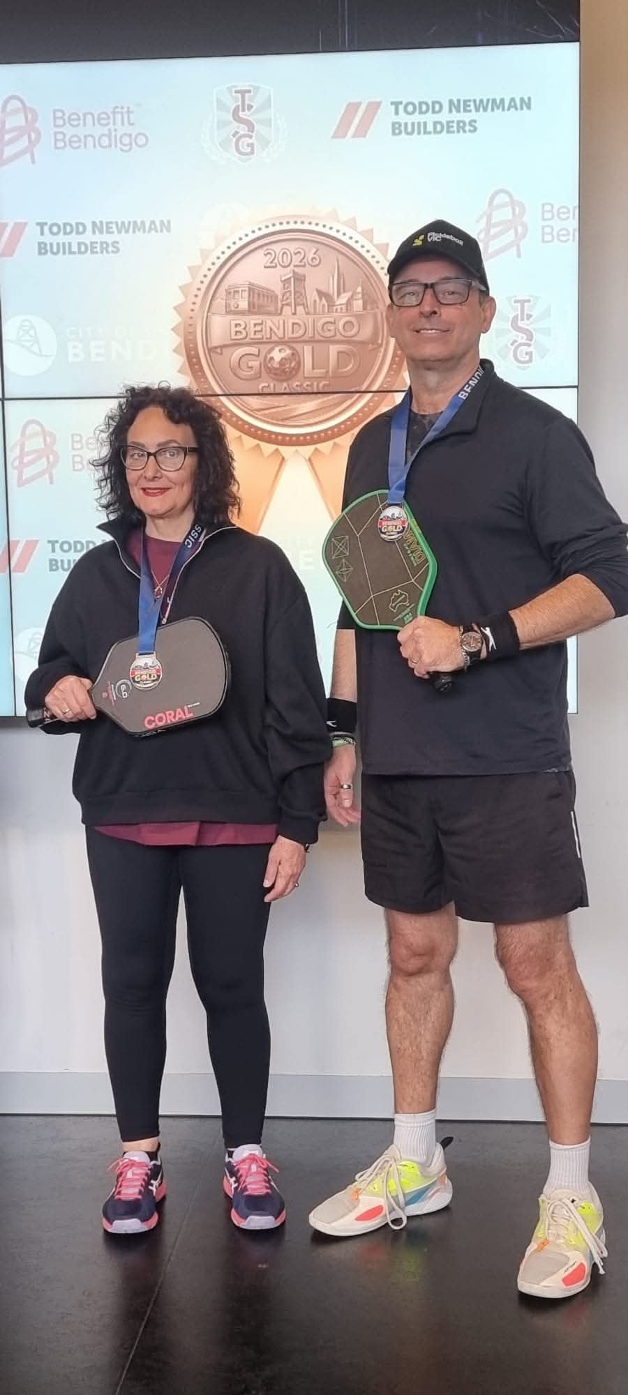 Two players with medals on grass beside outdoor pickleball courts
